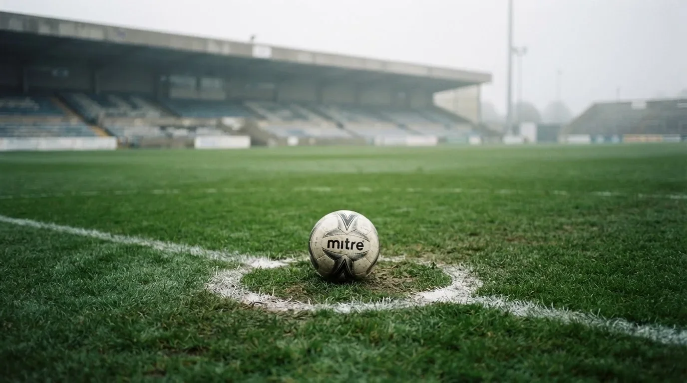 Ein Fußball liegt auf dem Elfmeterpunkt eines leeren Rasenplatzes, im Hintergrund eine Tribüne im Dunst.