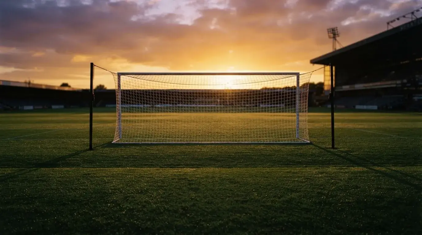 Ein Fußballtor mit Netz bei Sonnenuntergang auf einem leeren Platz, die Schatten der Torpfosten liegen lang auf dem Gras.