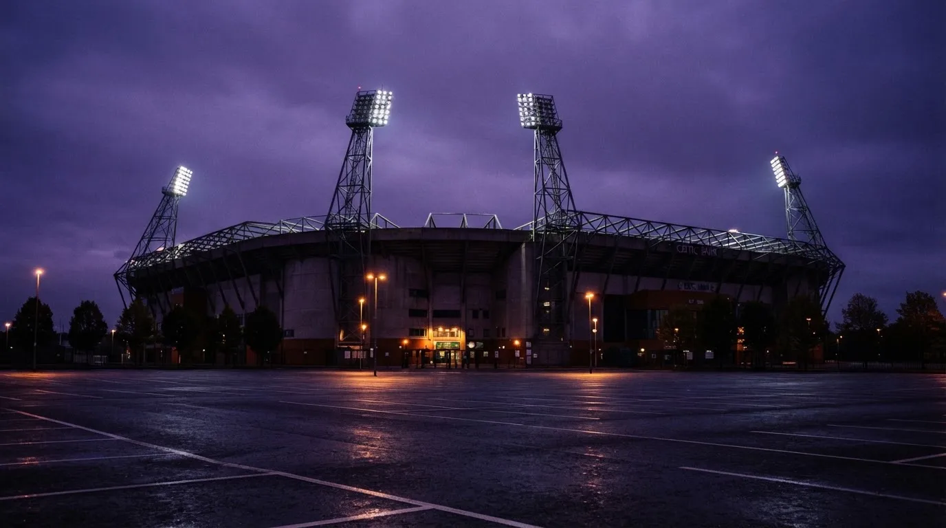 Ein Fußballstadion von außen in der Dämmerung, hohe Flutlichtmasten vor einem violetten Himmel, davor ein leerer Parkplatz.