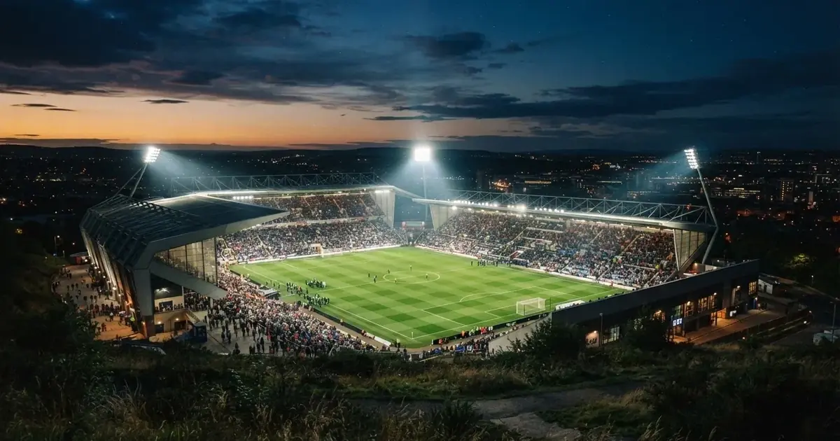 Ein Fußballstadion unter Flutlicht mit hellen Rängen, von einem Hügel aus gesehen, darüber ein dunkelblauer Abendhimmel.