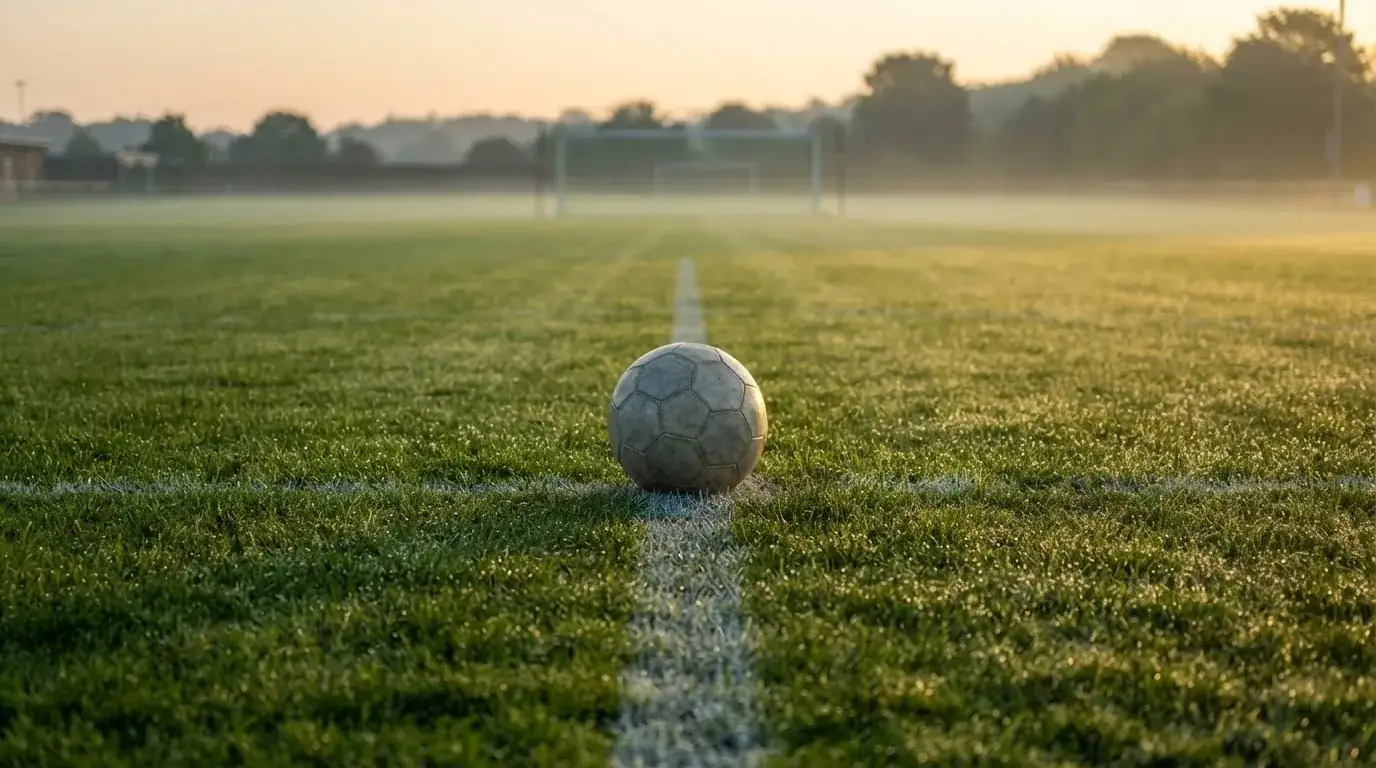 Ein Fußball liegt auf dem Anstoßpunkt eines Rasens bei Sonnenaufgang, Tau glitzert auf den Halmen.