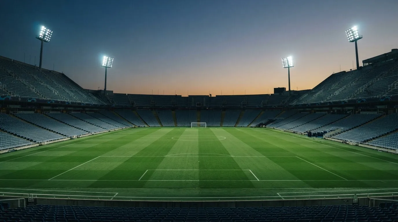 Eine leere Stadiontribüne unter Flutlicht aus der Ferne betrachtet, die Rasenfläche leuchtet grün in der Dämmerung.