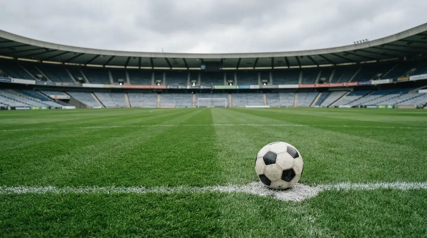 Ein Fußball liegt auf dem Anstoßpunkt eines leeren Rasenplatzes, im Hintergrund eine ruhige Tribüne unter bedecktem Himmel.