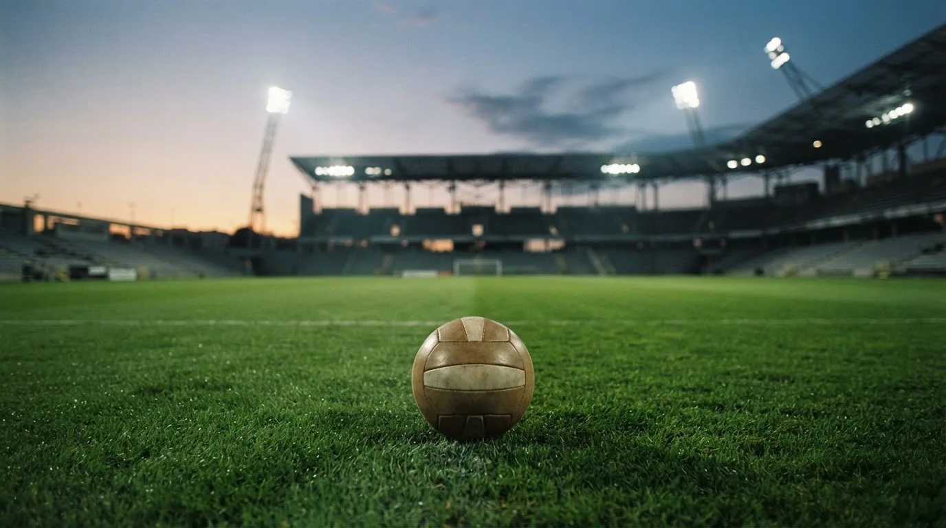 Ein Fußball liegt auf dem Rasen eines leeren Platzes, im Hintergrund ein Stadion unter Flutlicht in der Abenddämmerung.