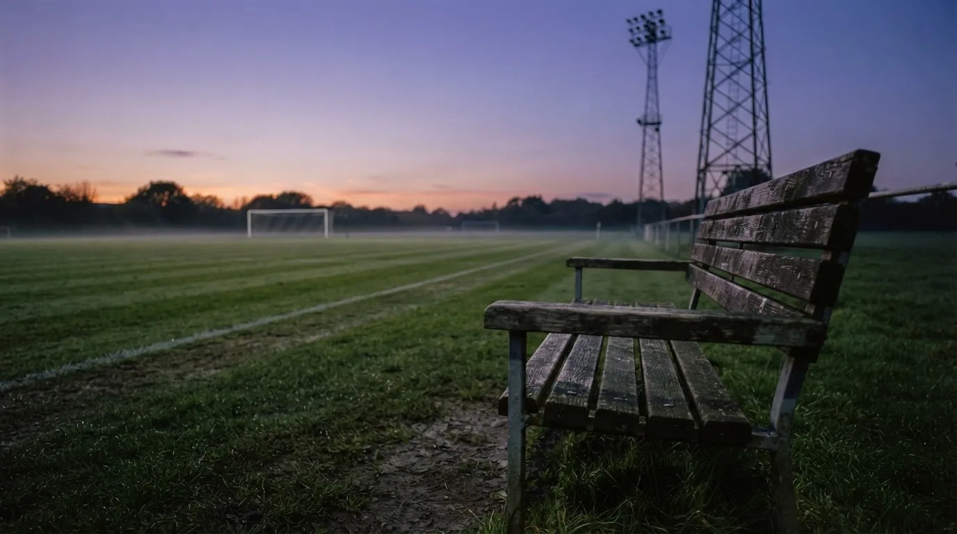 Eine leere Parkbank am Rand eines Fußballfelds in der Abenddämmerung, im Hintergrund ragen dunkle Flutlichtmasten in den Himmel.